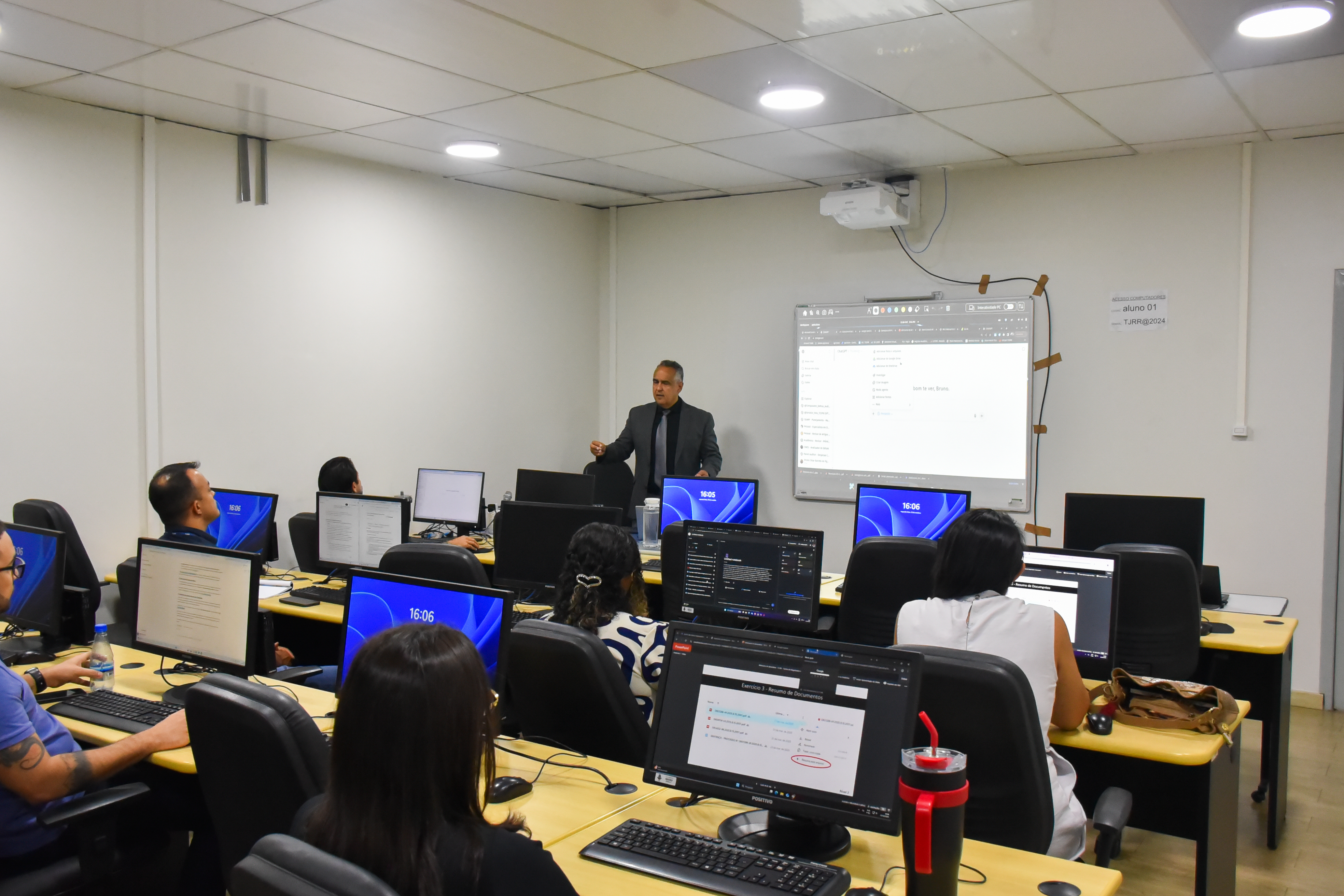 Imagem colorida mostra um ambiente de sala de aula ou laboratório de informática. Diversos alunos (homens e mulheres) estão sentados em mesas individuais com computadores, de costas ou de perfil para a câmera, concentrados em seus monitores. No centro da sala, um homem de terno cinza e gravata escura está de pé, gesticulando enquanto fala para a turma. Atrás dele, um projetor exibe informações em uma tela de projeção digital/quadro interativo. A tela mostra uma interface de computador com caixas de texto e dados. À direita da tela, em um quadro branco, há anotações simples. O ambiente é claro, com iluminação de teto. Muitos dos monitores estão exibindo uma tela azul brilhante, alguns exibem formulários ou documentos. Imagem colorida mostra um ambiente de sala de aula ou laboratório de informática. Diversos alunos (homens e mulheres) estão sentados em mesas individuais com computadores, de costas ou de perfil para a câmera, concentrados em seus monitores. No centro da sala, um homem de terno cinza e gravata escura está de pé, gesticulando enquanto fala para a turma. Atrás dele, um projetor exibe informações em uma tela de projeção digital/quadro interativo. A tela mostra uma interface de computador com caixas de texto e dados. À direita da tela, em um quadro branco, há anotações simples. O ambiente é claro, com iluminação de teto. Muitos dos monitores estão exibindo uma tela azul brilhante, alguns exibem formulários ou documentos.