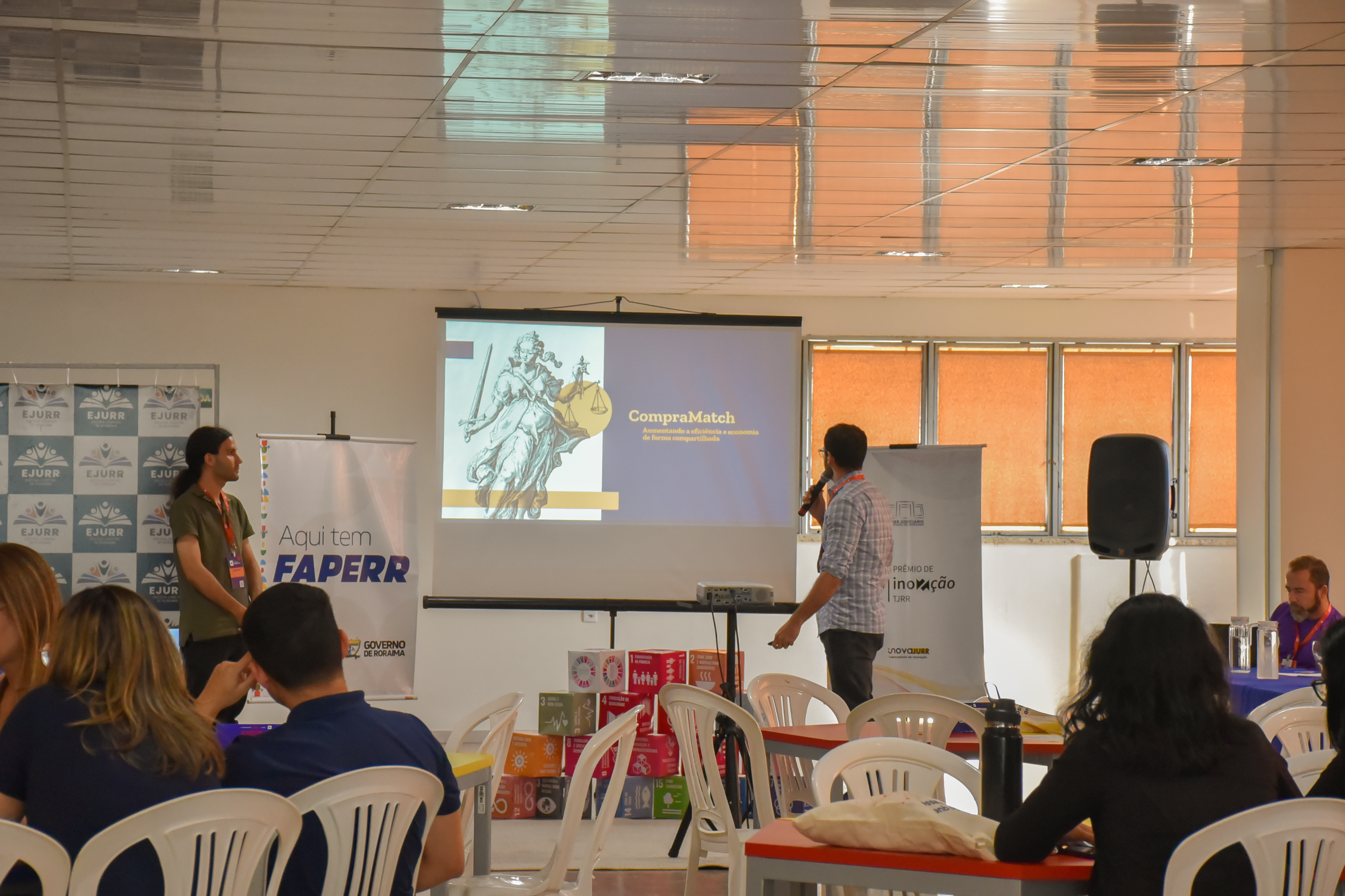 Imagem colorida mostra um evento ou workshop em um salão iluminado. A câmera captura um momento de apresentação. No centro, um homem com camisa quadriculada, de costas para a plateia, está falando em um microfone. Atrás dele, um telão de projeção exibe um slide com o título "CompraMatch" e a imagem de uma figura feminina clássica (possivelmente a deusa da justiça) ao lado de um leão. Há banners com logos, incluindo o da EJURR e da FAPERR, ao lado da tela. Na frente, duas mulheres e um homem estão sentados em cadeiras brancas de plástico em mesas vermelhas e amarelas, de perfil ou de costas para a câmera, interagindo ou assistindo à apresentação. Caixas com logos dos Objetivos de Desenvolvimento Sustentável (ODS) da ONU estão empilhadas no chão, perto da mesa. O teto é de laje e o ambiente é bem iluminado, com janelas grandes cobertas por persianas laranjas no fundo. Imagem colorida mostra um evento ou workshop em um salão iluminado. A câmera captura um momento de apresentação. No centro, um homem com camisa quadriculada, de costas para a plateia, está falando em um microfone. Atrás dele, um telão de projeção exibe um slide com o título "CompraMatch" e a imagem de uma figura feminina clássica (possivelmente a deusa da justiça) ao lado de um leão. Há banners com logos, incluindo o da EJURR e da FAPERR, ao lado da tela. Na frente, duas mulheres e um homem estão sentados em cadeiras brancas de plástico em mesas vermelhas e amarelas, de perfil ou de costas para a câmera, interagindo ou assistindo à apresentação. Caixas com logos dos Objetivos de Desenvolvimento Sustentável (ODS) da ONU estão empilhadas no chão, perto da mesa. O teto é de laje e o ambiente é bem iluminado, com janelas grandes cobertas por persianas laranjas no fundo.