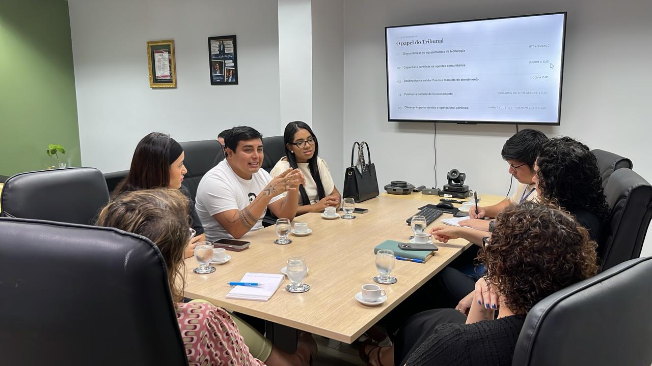 Reunião em uma sala, com sete pessoas sentadas em torno de uma mesa, discutindo. Um telão exibe informações sobre o papel do Tribunal. Há copos de água e xícaras de café na mesa.