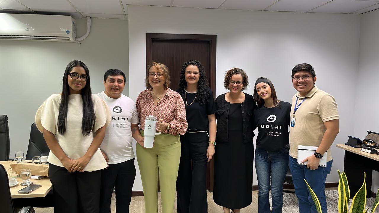 Sete pessoas estão sorrindo lado a lado em uma sala de reuniões comercial, com duas delas vestindo camisetas pretas e brancas da Associação Yanomami Urihi.