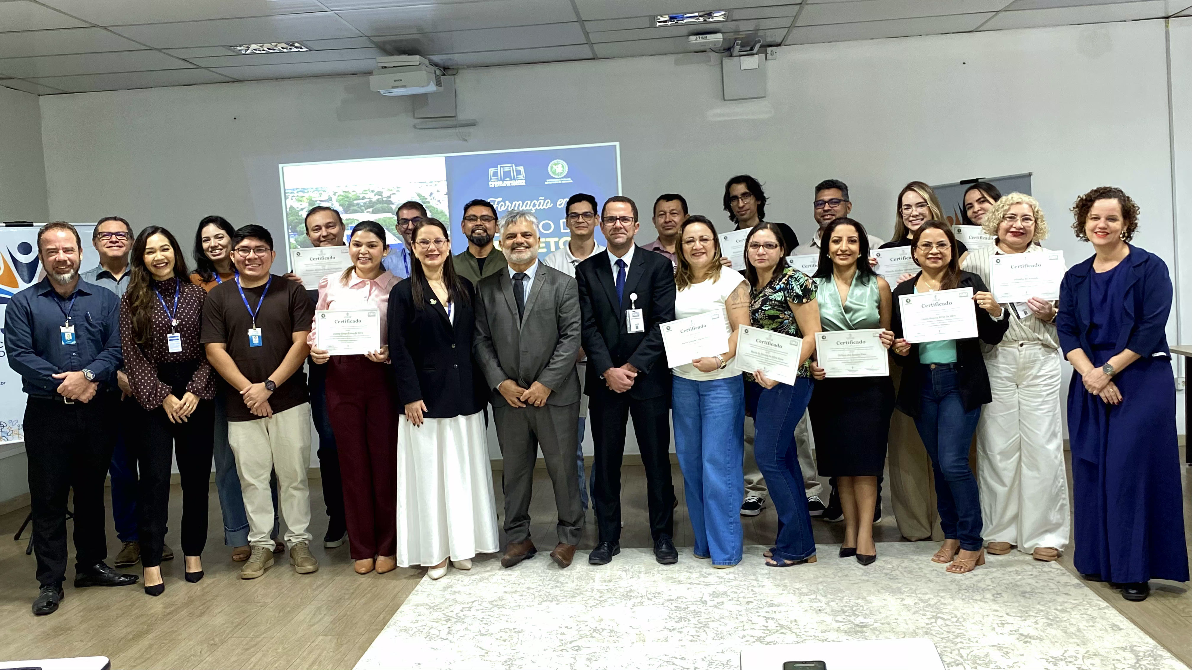 Foto em grupo dos participantes reunidos lado a lado. Eles sorriem para a câmera e vários seguram certificados brancos nas mãos. Foto em grupo dos participantes reunidos lado a lado. Eles sorriem para a câmera e vários seguram certificados brancos nas mãos.