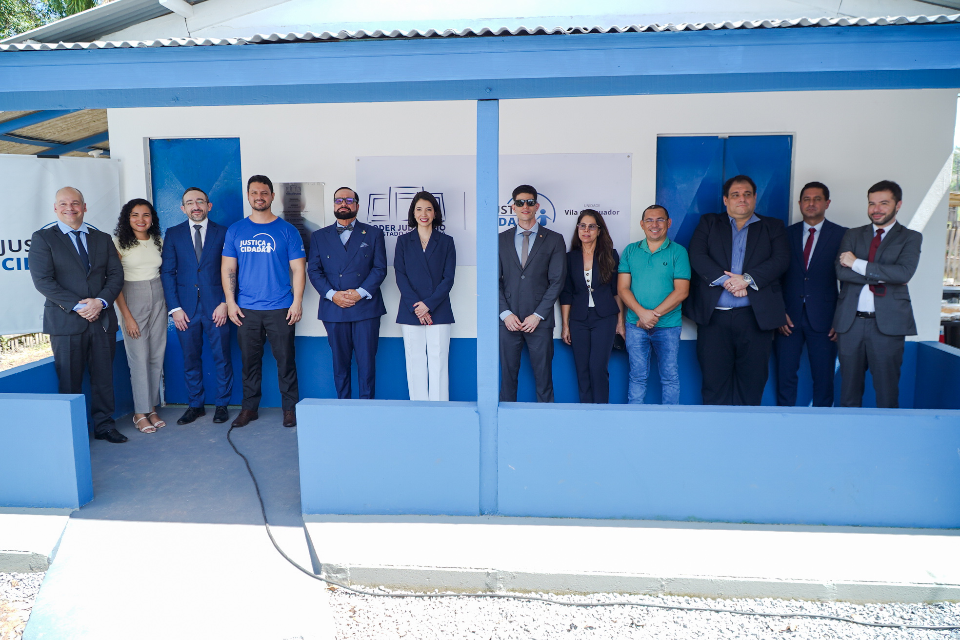 Fotografia colorida registra magistrados, representantes de instituições e autoridades posando para foto oficial diante da Unidade Justiça Cidadã da Vila do Equador. Ao fundo aparecem a fachada azul e branca e as placas de identificação do novo espaço.