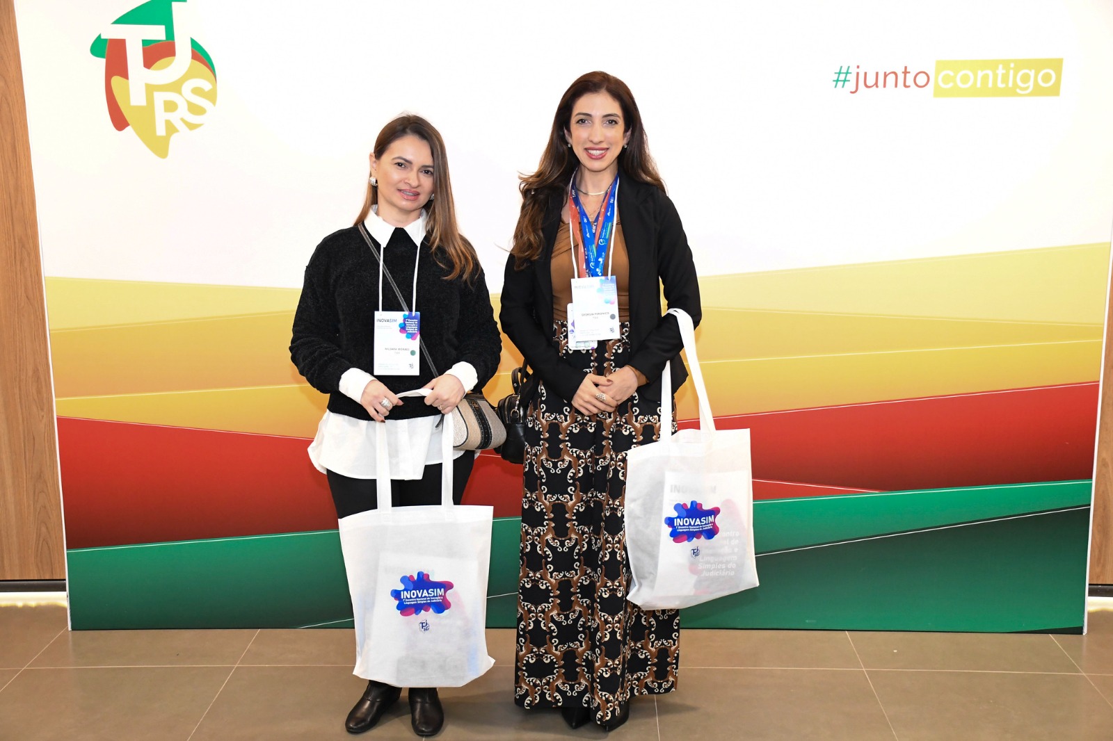 Foto 3: Imagem colorida mostra duas mulheres posadas lado a lado em frente a um painel institucional. Ambas usam crachás do evento e seguram sacolas ecológicas brancas com o logotipo "INOVASIM". À esquerda, uma mulher veste camisola preta sobre camisa branca com gola à mostra; à direita, a outra mulher veste blazer preto, blusa castanha e calça estampada. O painel ao fundo traz as cores verde, amarela e vermelha, o logotipo do TJRS e a hashtag "#juntocontigo". Fim da descrição. Foto 3: Imagem colorida mostra duas mulheres posadas lado a lado em frente a um painel institucional. Ambas usam crachás do evento e seguram sacolas ecológicas brancas com o logotipo "INOVASIM". À esquerda, uma mulher veste camisola preta sobre camisa branca com gola à mostra; à direita, a outra mulher veste blazer preto, blusa castanha e calça estampada. O painel ao fundo traz as cores verde, amarela e vermelha, o logotipo do TJRS e a hashtag "#juntocontigo". Fim da descrição.