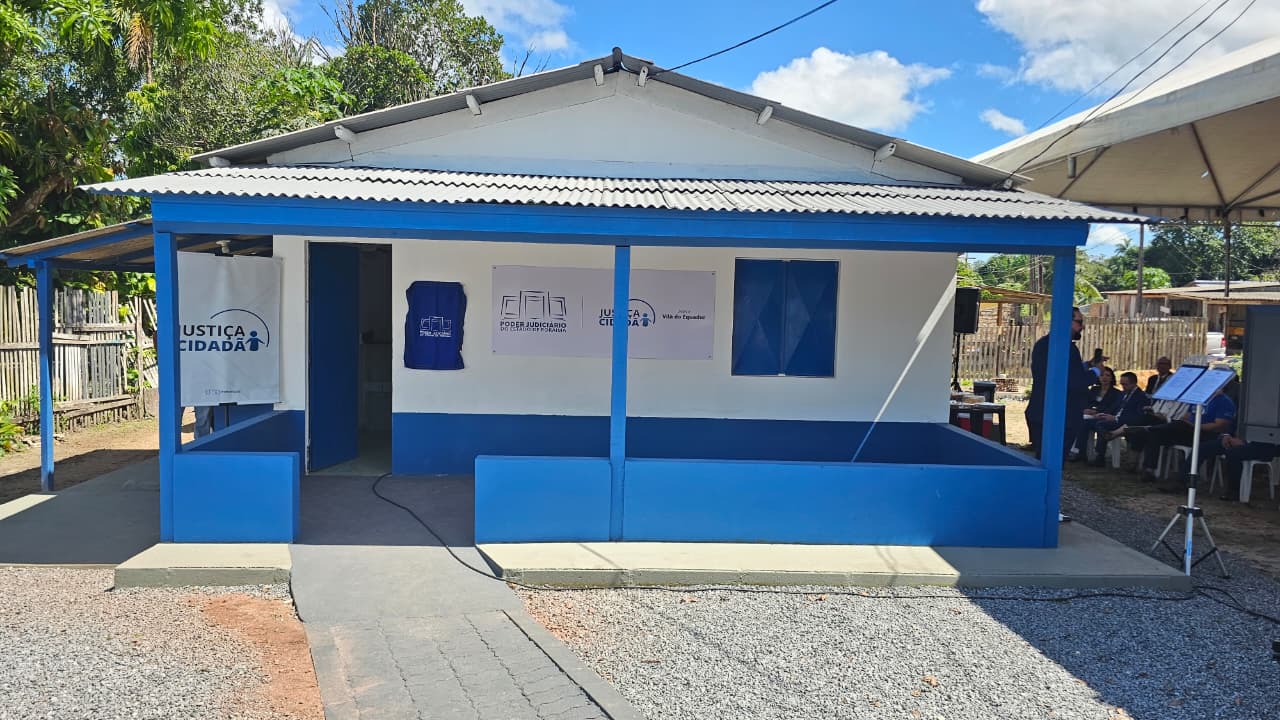 a fachada da unidade, pintada nas cores branca e azul, com as placas de identificação do Poder Judiciário de Roraima e do programa Justiça Cidadã. O espaço é simples, com varanda frontal e acesso por uma calçada de concreto. Ao redor, há árvores e residências da comunidade.